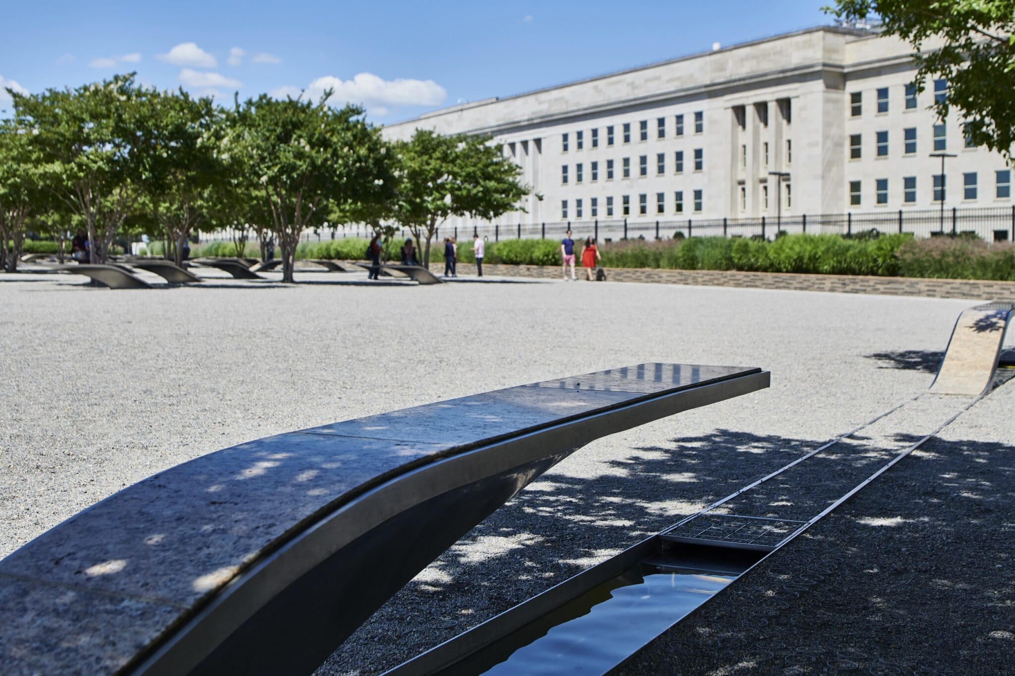 Memorial with curved benches over small reflecting pools, set in a gravel area with trees in the background. A large building is visible under a clear blue sky. People are walking in the distance.