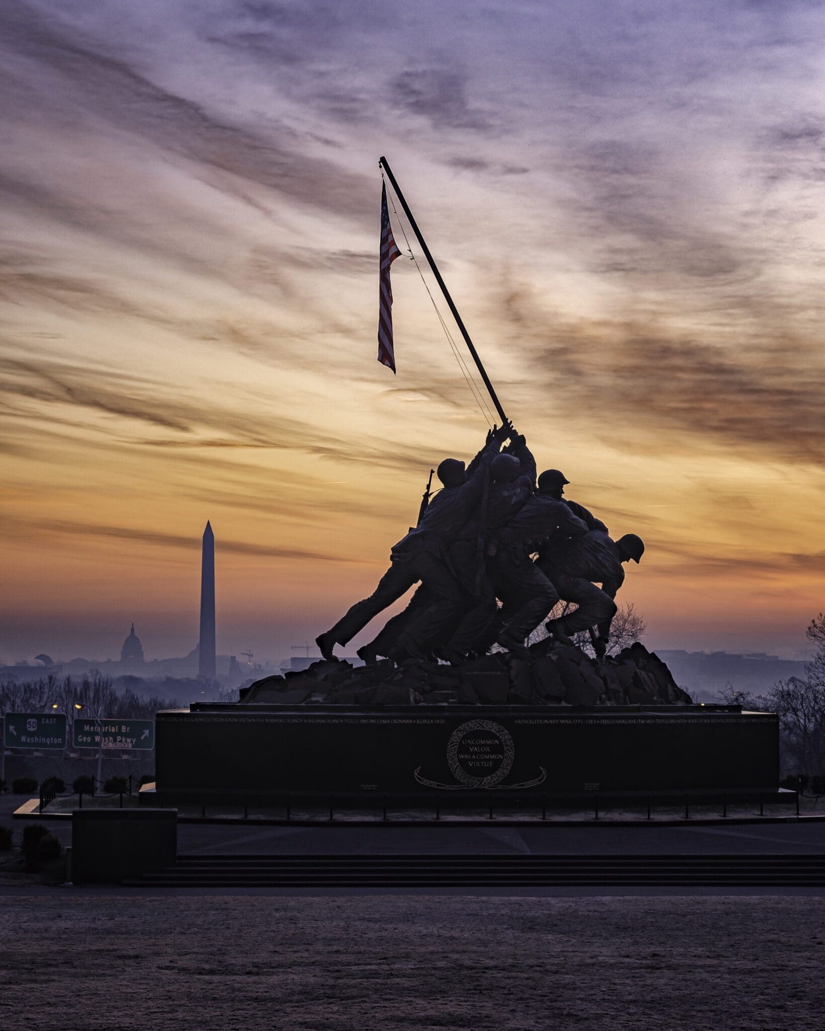 Silhouetted Iwo Jima Memorial with soldiers raising a flag at sunrise. The Washington Monument and U.S. Capitol are visible in the hazy background against a sky filled with orange and purple hues.
