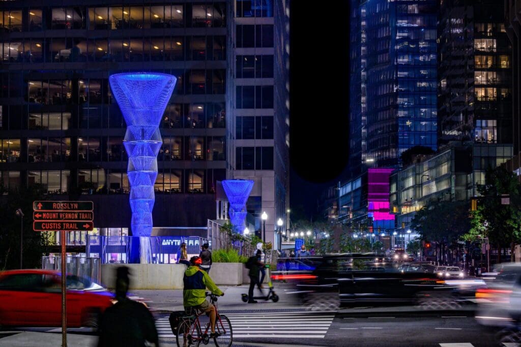A vibrant city street at night with illuminated buildings and two large cone-shaped light sculptures. Cyclists and scooters are in motion, and cars blur in the foreground, showcasing the hustle and bustle of urban life.