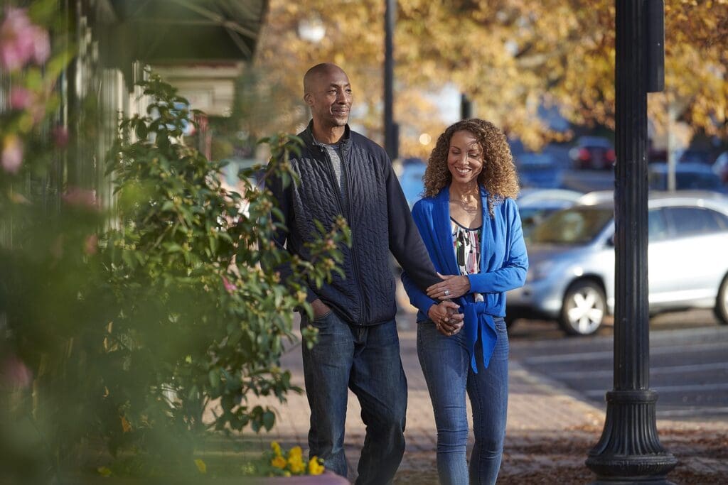 A couple walks hand in hand on a sidewalk lined with greenery. They are smiling and enjoying the autumn day, with colorful leaves in the background and parked cars nearby. The man is wearing a dark sweater, and the woman is in a blue cardigan.