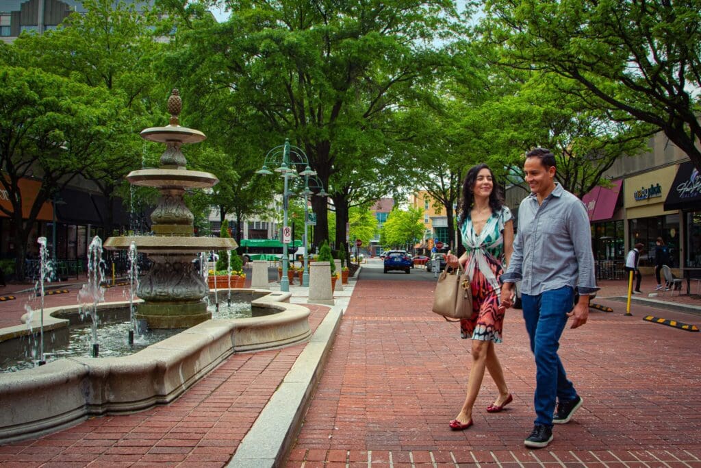 A couple walks hand in hand on a brick-paved street lined with trees. A decorative fountain is on their left. The street has shops and parked cars, and the atmosphere is vibrant and relaxed.