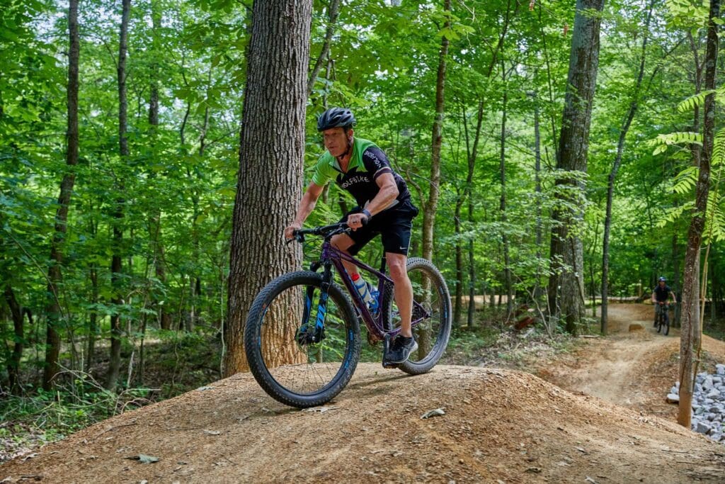 A person wearing a helmet and cycling gear is riding a mountain bike on a dirt trail in a forest. The trail features a small hill. Another cyclist is visible in the background. Tall trees and green foliage surround the path.
