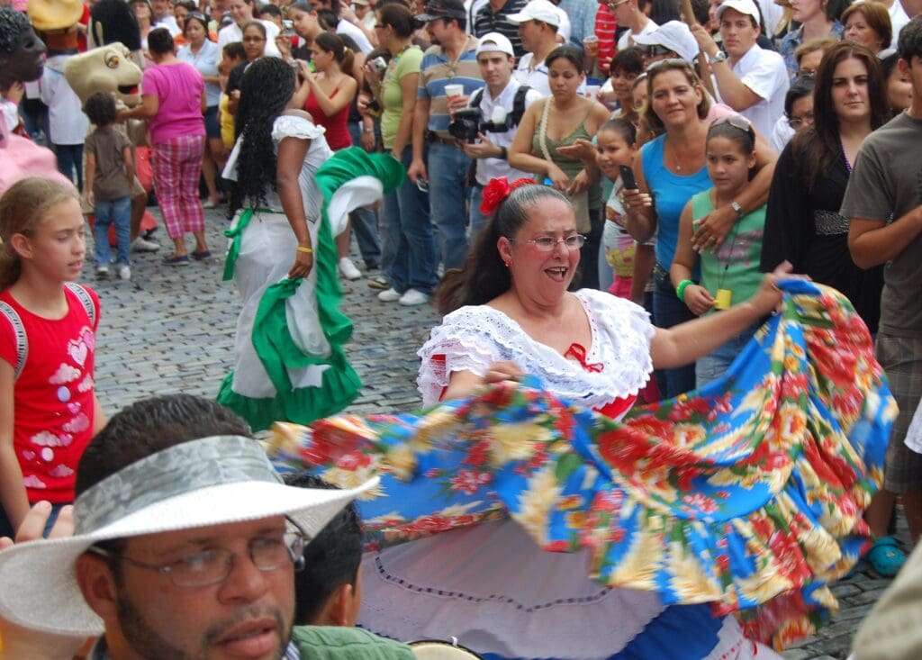 A woman in festive, colorful traditional attire dances joyfully among a crowd on a cobblestone street. She swings her vibrant patterned skirt. Onlookers, including adults and children, gather around, some taking photos. The atmosphere is lively and celebratory.