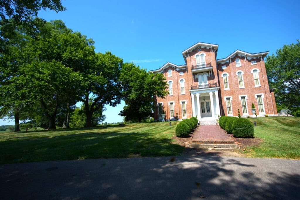 A large, historic brick house with white trim and tall windows under a clear blue sky. It is surrounded by lush green trees and a well-kept lawn, with a brick pathway leading to the entrance.
