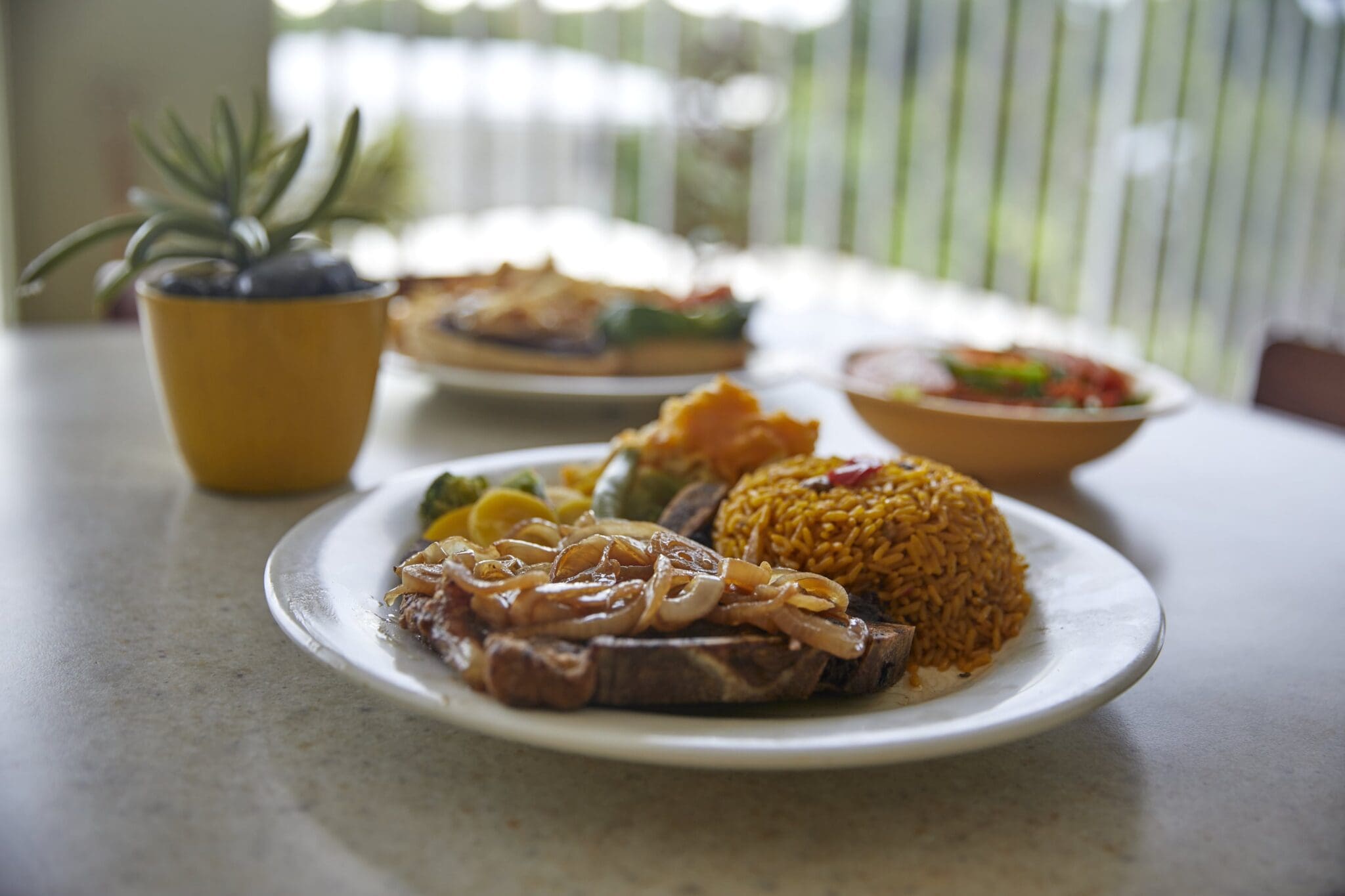 A plate with beef topped with sautéed onions, accompanied by seasoned rice, grilled vegetables, and a slice of lemon. A small plant and another bowl of food are blurred in the background on a table.