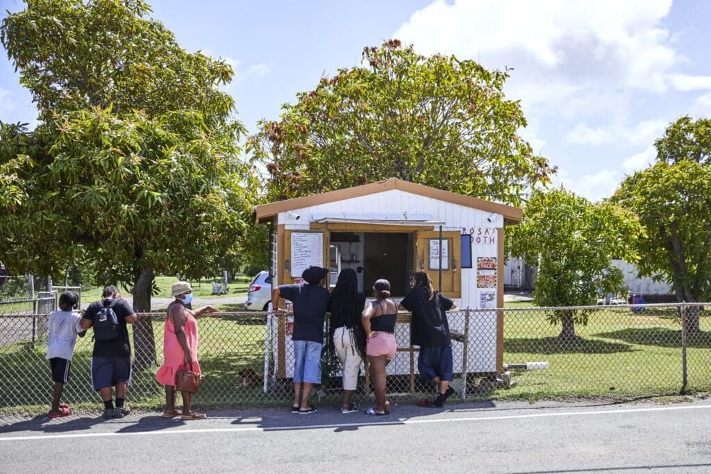 People stand in line at a small roadside food stall with a menu posted outside. The stall is surrounded by green trees and enclosed by a chain-link fence. A car is parked in the background on a sunny day.