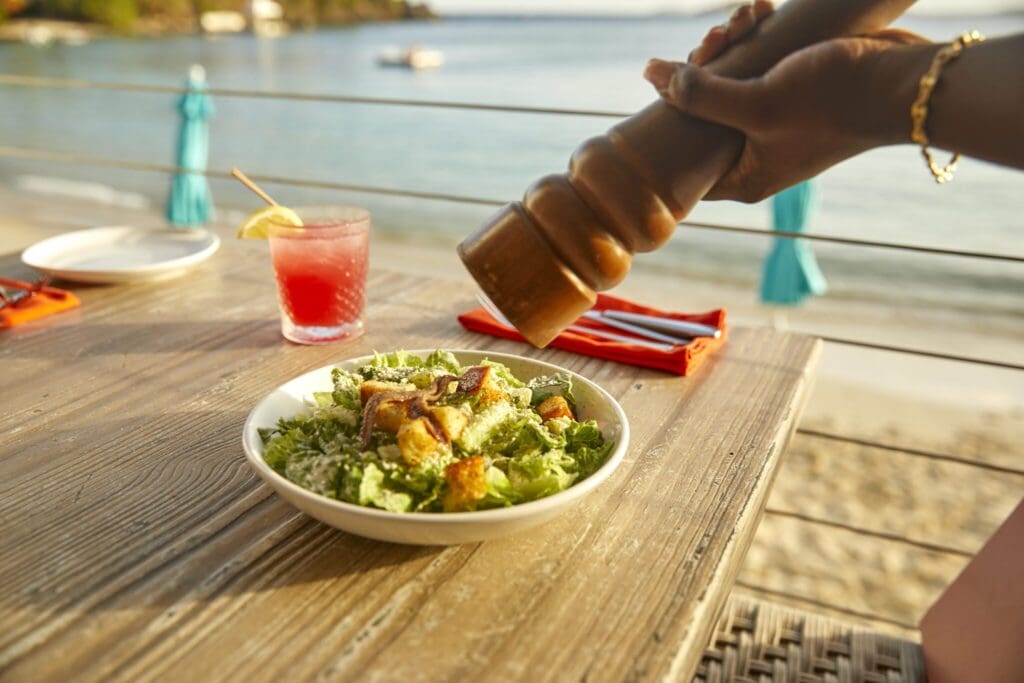 A close-up of a persons hand using a large pepper grinder over a Caesar salad on a wooden table. Theres a red drink with a lemon slice, a plate, and a sea view in the background. The table is set with orange napkins and utensils.