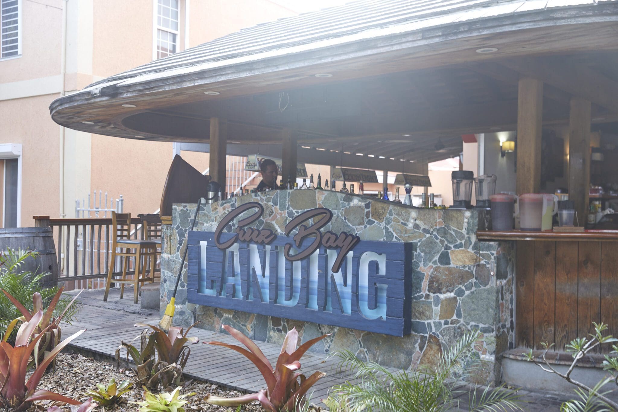 Outdoor bar with a rustic stone and wood design. A large sign reads Cruz Bay Landing. Surrounded by plants and featuring a roofed seating area.