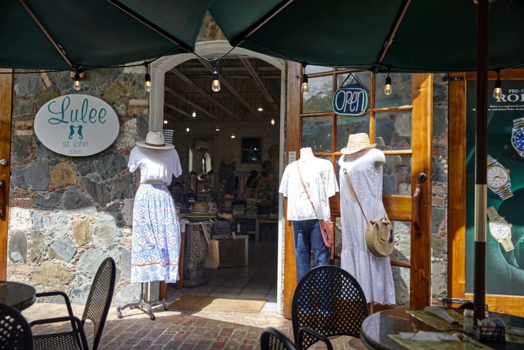 Entrance to a boutique named Lulee displaying mannequins in summer dresses and hats outside. A sign reads Open. The interior is visible through wooden-framed glass doors. Tables and chairs are in the foreground under a green umbrella.