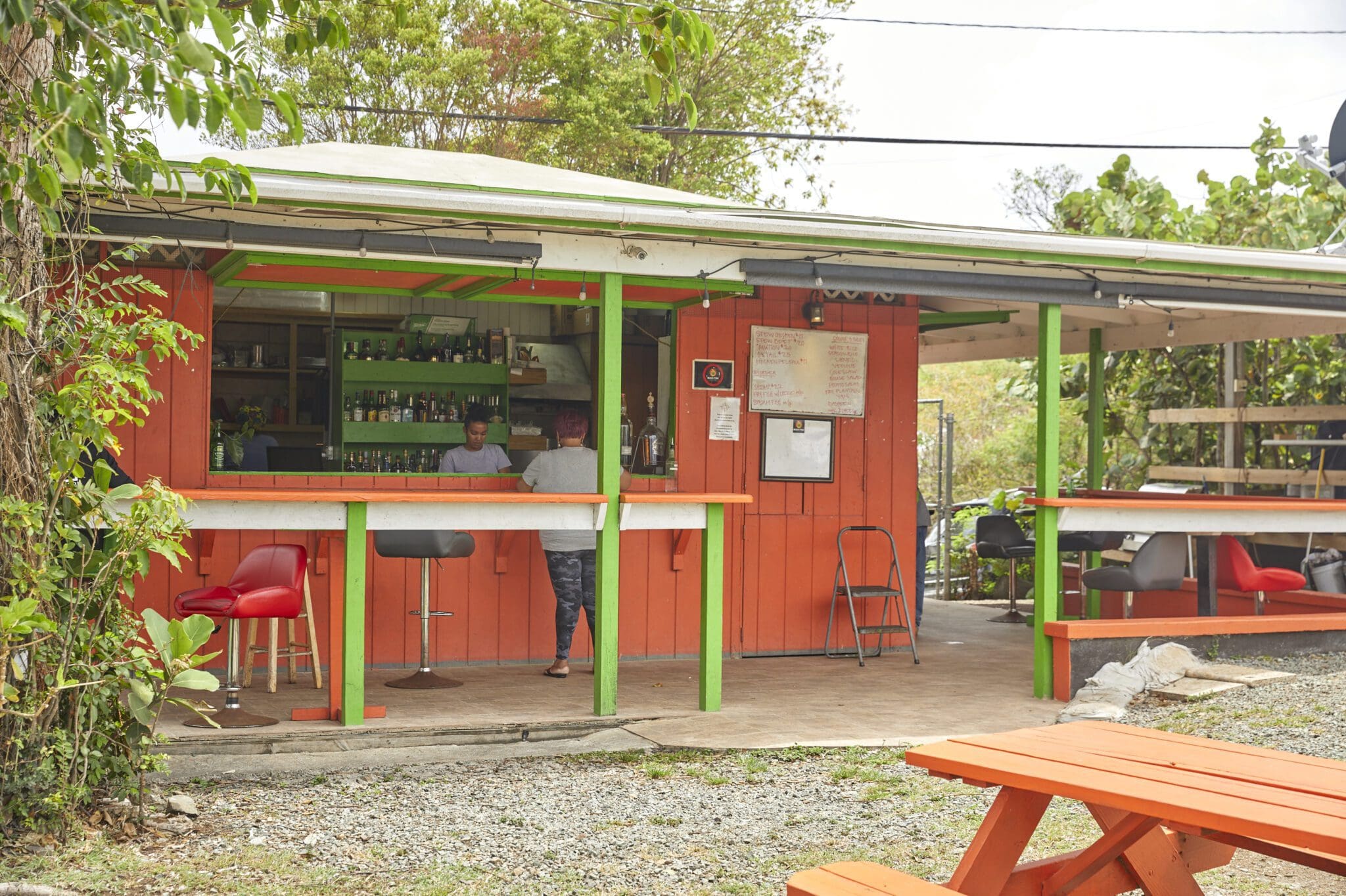 An outdoor bar with bright orange and green accents. Two people are sitting on bar stools, facing the bar. A picnic table sits in the foreground. The area is surrounded by greenery. The menu is displayed on the wall behind the bar.