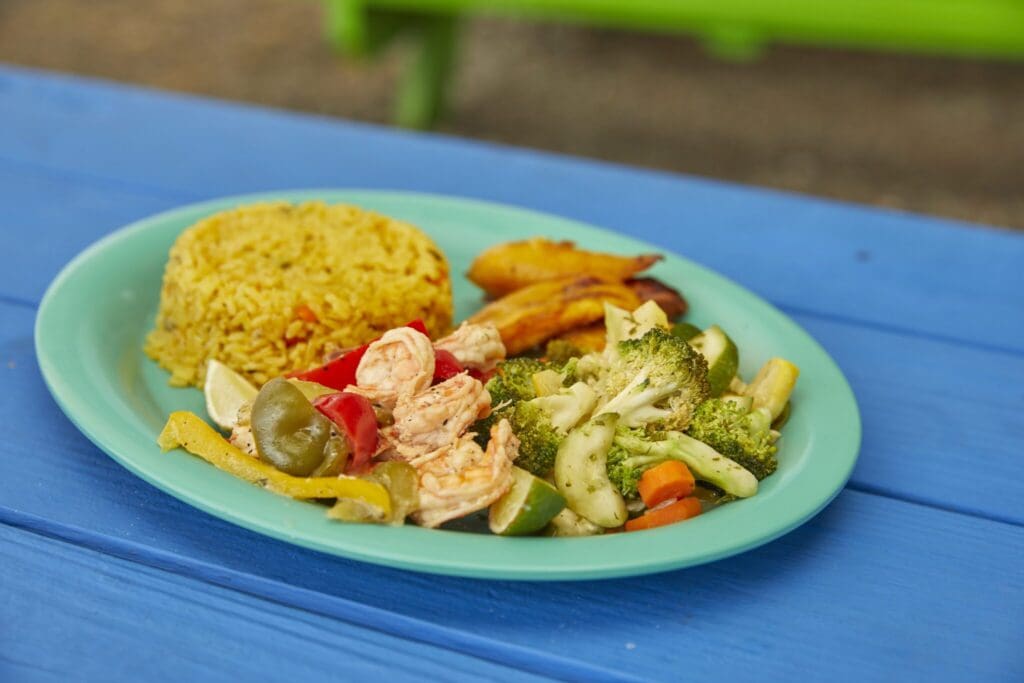 A turquoise plate on a blue table holds yellow rice, sautéed mixed vegetables, including broccoli and peppers, cooked shrimp, and fried plantains. The setting appears outdoor with a green background.