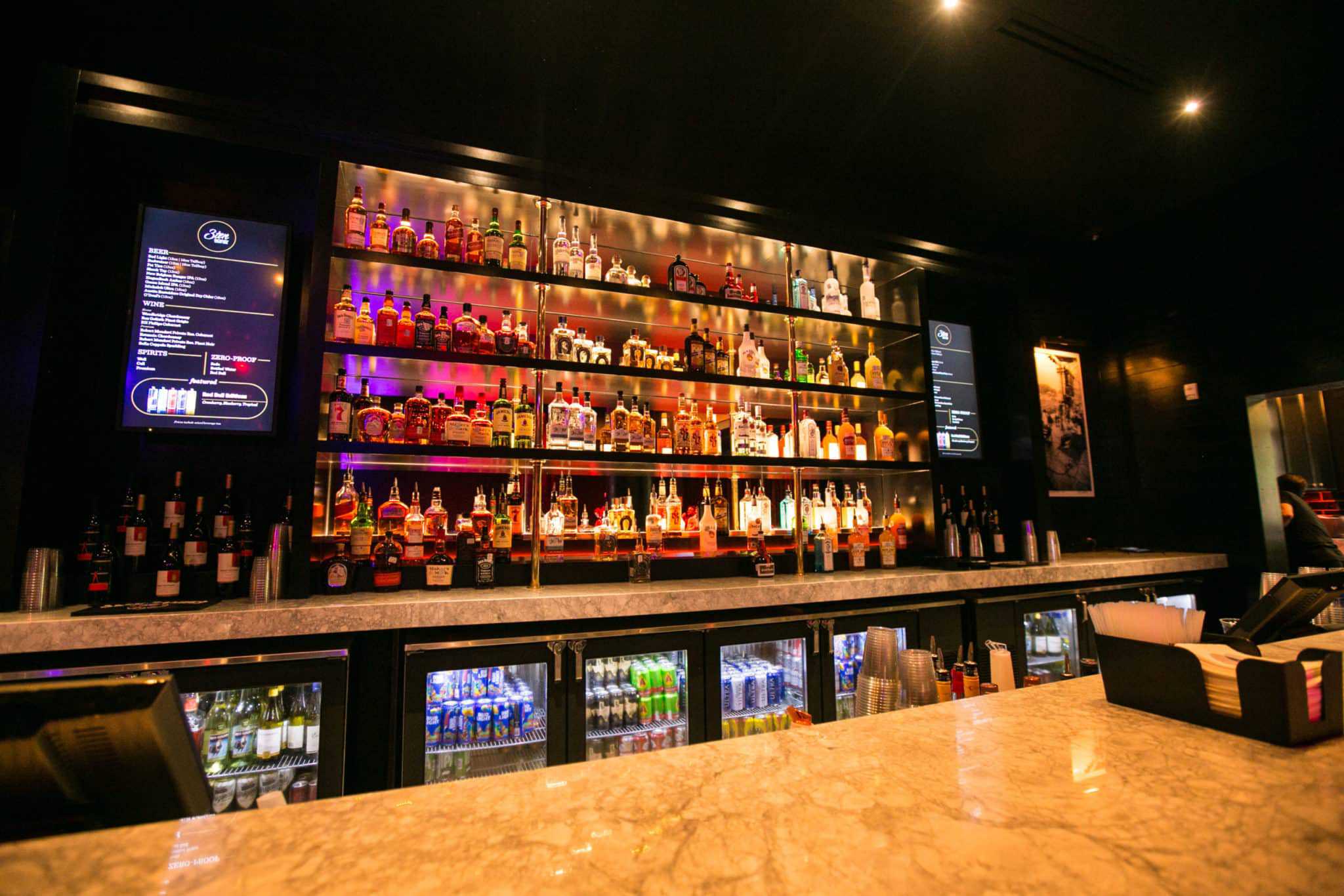 A dimly lit bar with a marble countertop and shelves stocked with various liquor bottles. Two digital screens display drink menus on either side of the shelves. A cooler with beverages is underneath the counter.