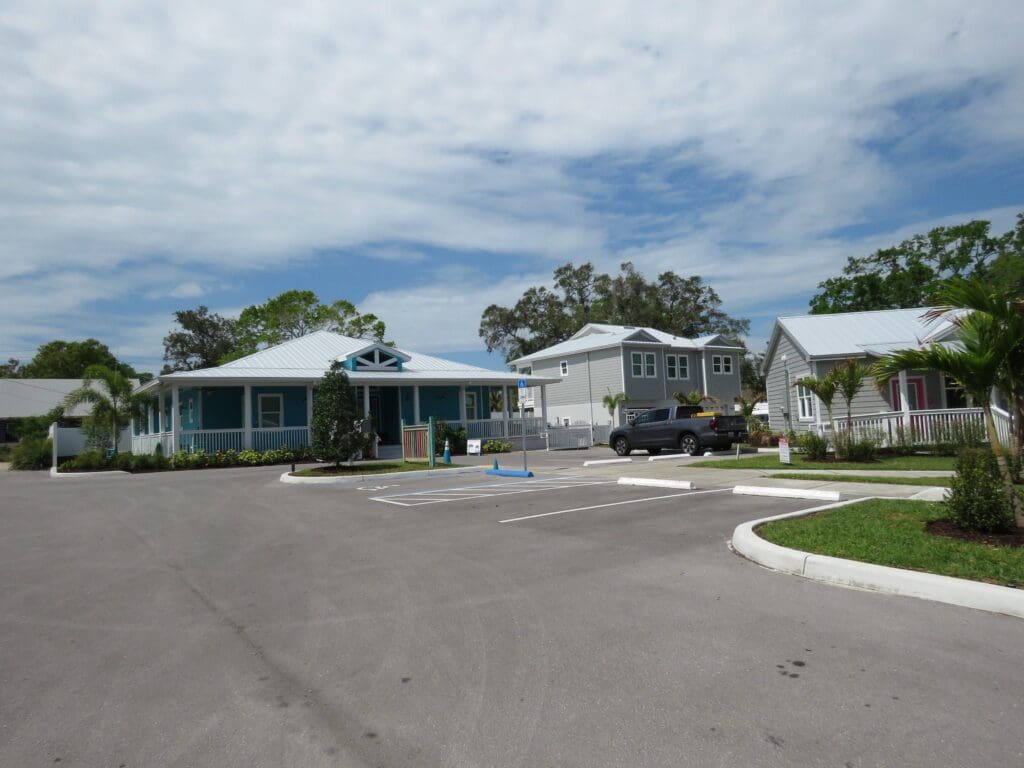 Wide shot of a parking lot in a residential area, featuring a blue house on the left and a gray house on the right. A pickup truck is parked near the houses. The sky is partly cloudy, and trees and greenery surround the area.
