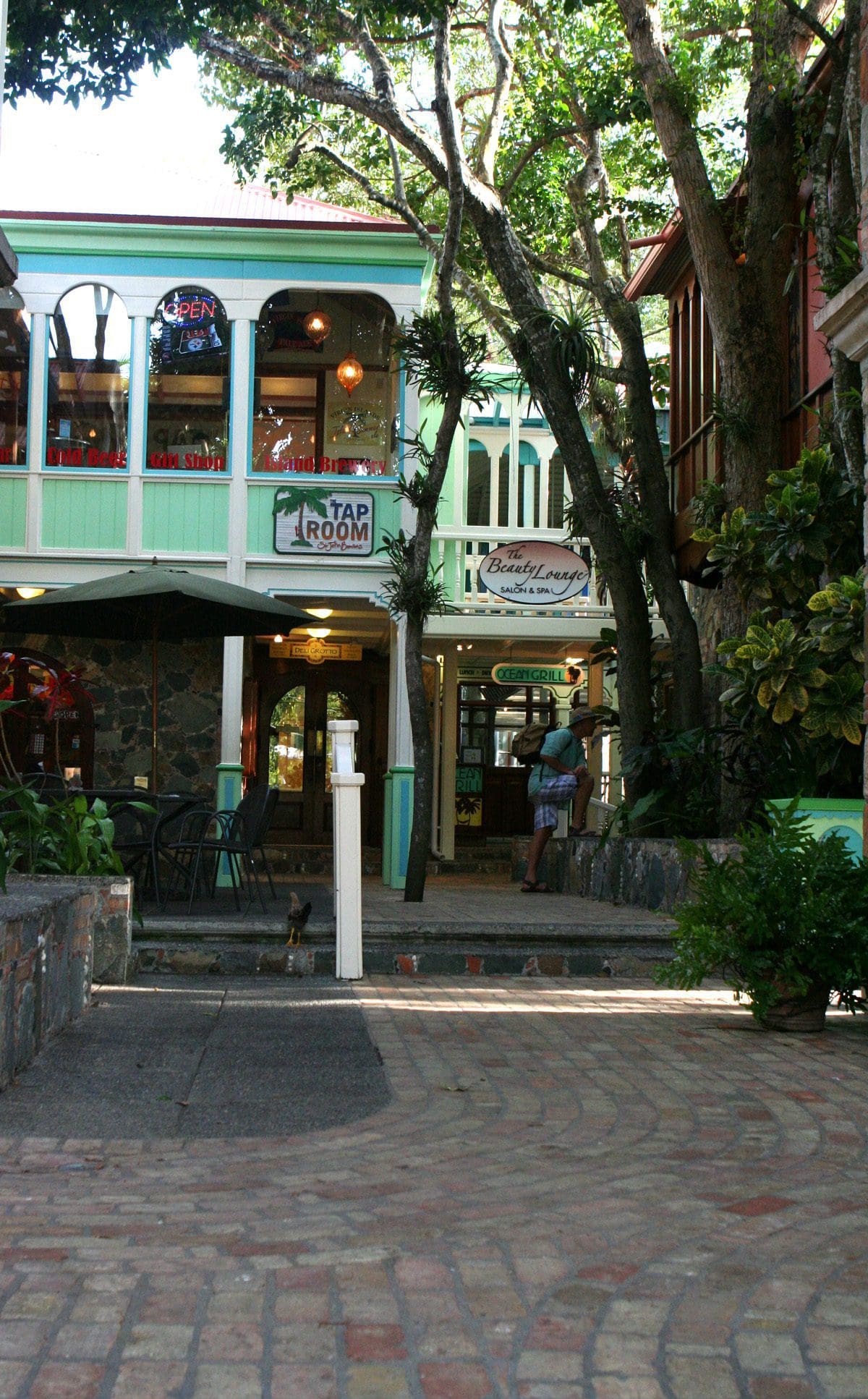 Shaded courtyard with brick path leading to a two-story building with a sign reading Tap Room. Trees surround the area, providing shade. An open sign is visible in one of the windows, and a person stands near the entrance.