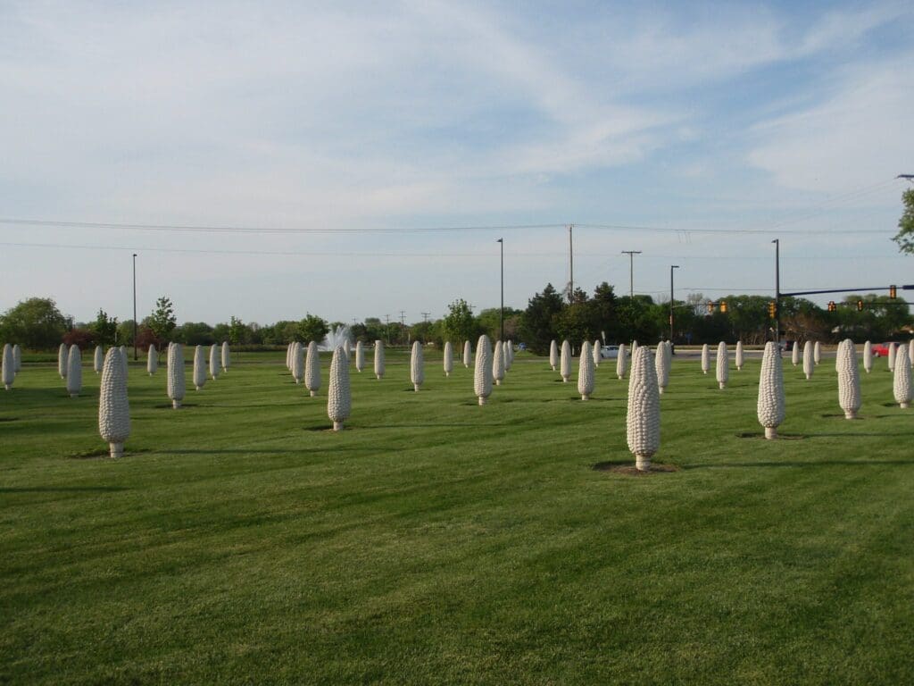 A field with numerous large, white, corn cob sculptures standing upright on a grassy lawn. The sky is mostly clear with a few clouds. Trees and streetlights are visible in the background.