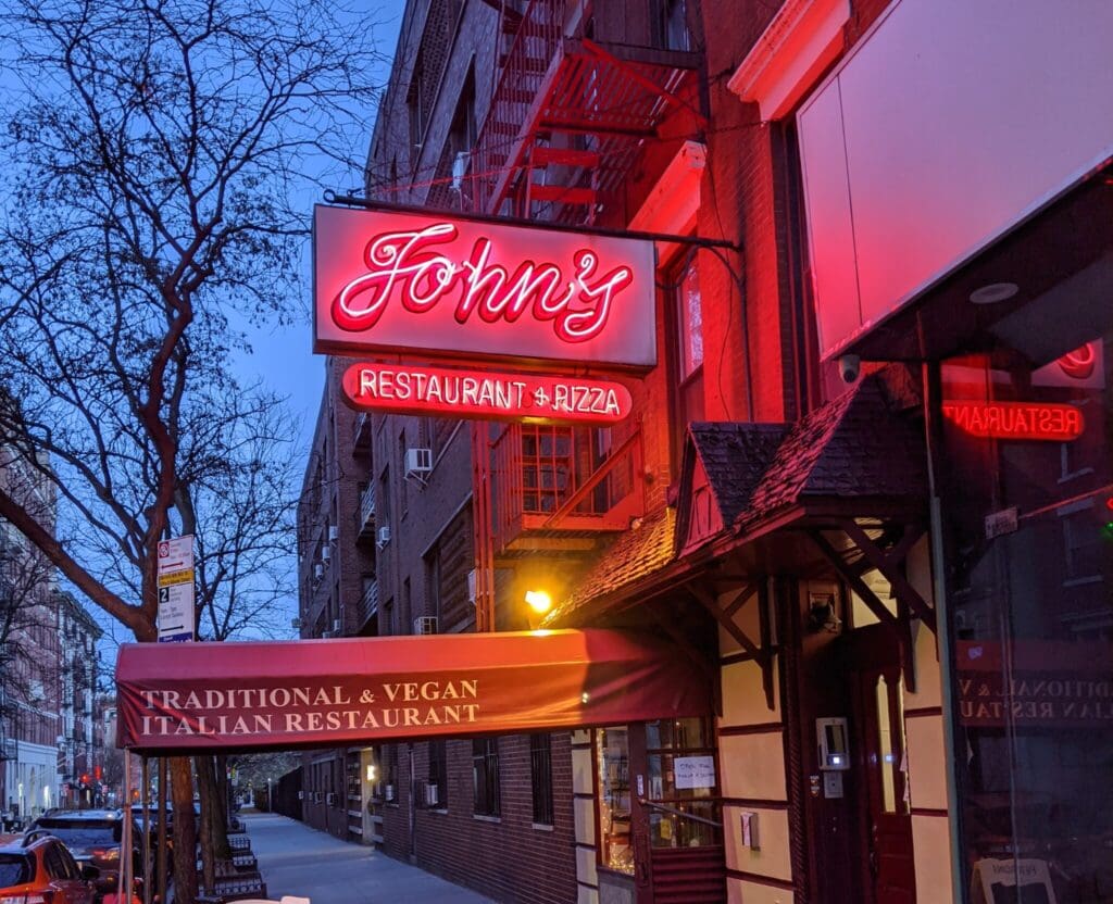 A city street scene with a red neon sign reading Johnnys Restaurant & Pizza on a brick building. Below, an awning displays Traditional & Vegan Italian Restaurant. Bare trees line the street, and the sidewalk is empty.