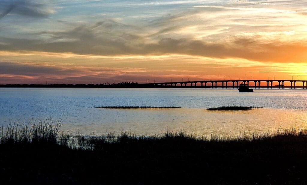 A serene sunset view over a body of water, with wispy clouds in the sky. A bridge stretches across the horizon, and a boat is visible in the distance. Grasses line the edge of the water in the foreground.