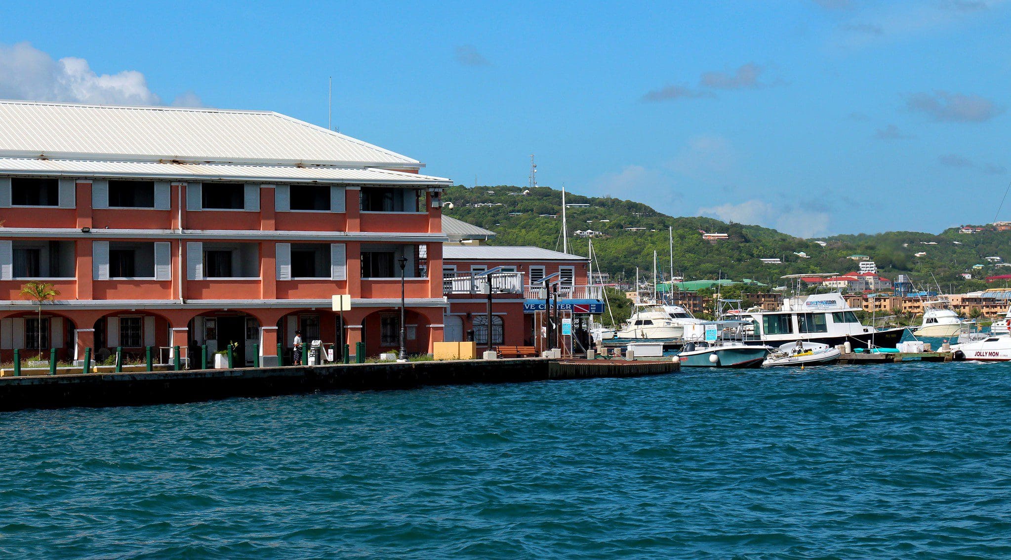 A marina with several boats docked near a red and white multi-story building. The ocean water is turquoise, and the backdrop features lush green hills and a blue sky with a few clouds.