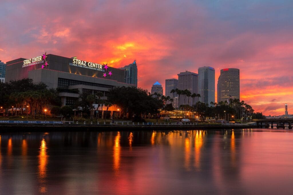 Sunset view of a city skyline with tall buildings, including the Straz Center, reflecting on a calm river. The sky is vibrant with hues of orange, pink, and purple. Silhouetted trees and a bridge are visible in the background.