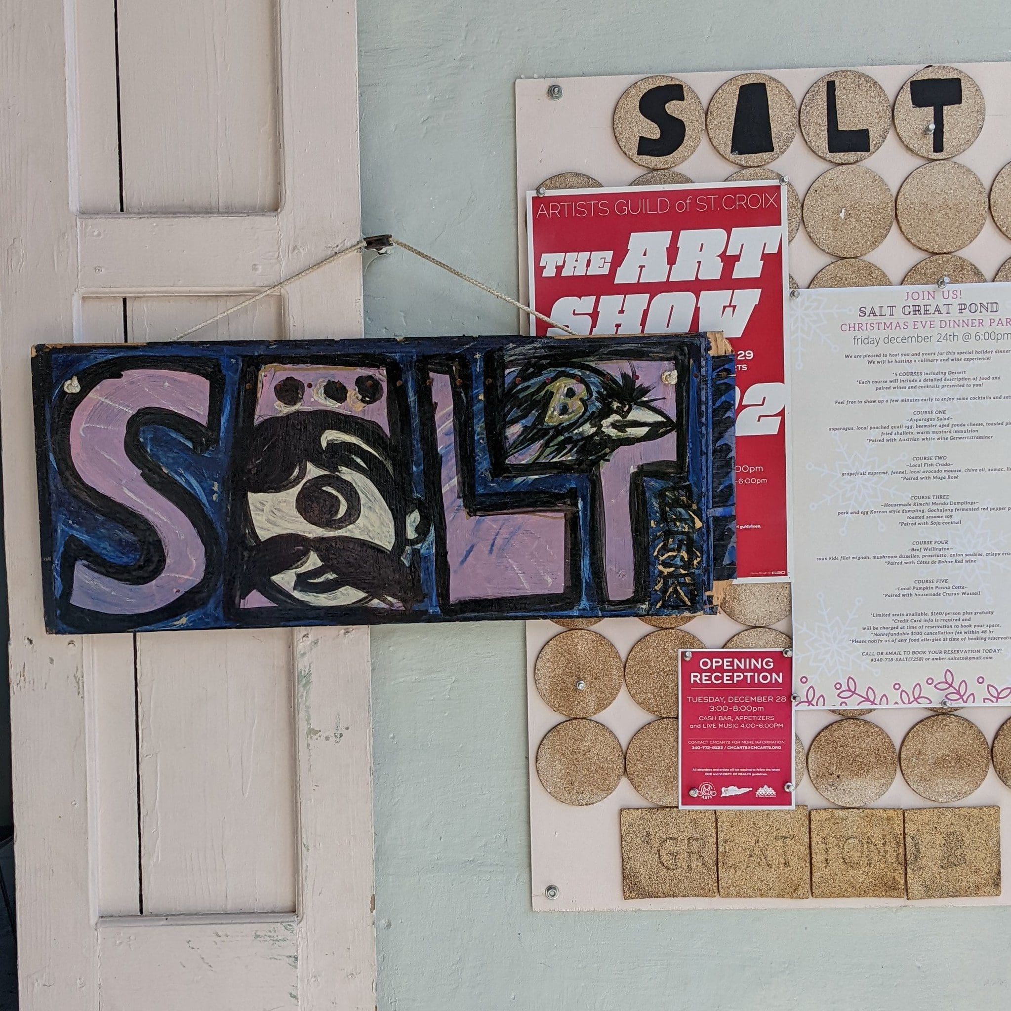 A black and purple sign with the word SALT in bold letters is hanging on a wall. Behind it, theres a bulletin board with multiple circular notices and a white paper listing event details. The area appears to be an art or community space.