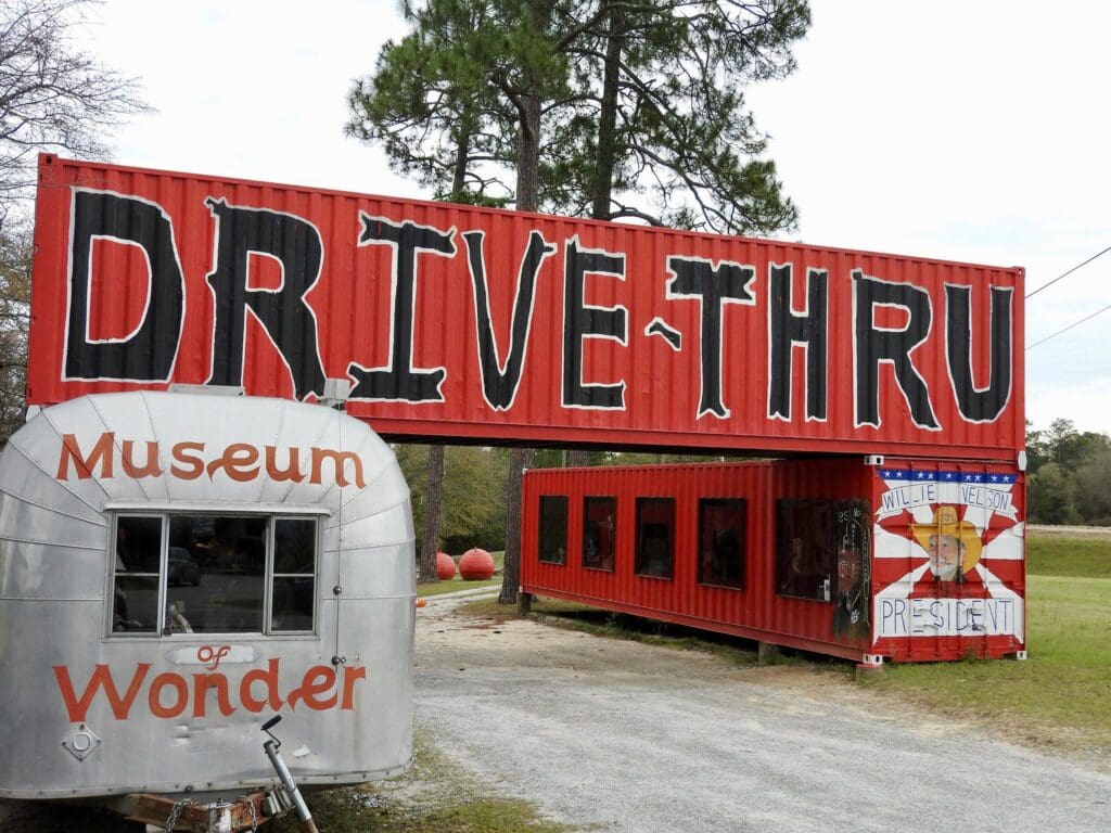 A large red shipping container sign reading DRIVE-THRU spans the entrance to a gravel driveway. Nearby, a silver trailer marked Museum of Wonder is seen. A poster with a caricature and the text WILLIE NELSON FOR PRESIDENT is displayed.