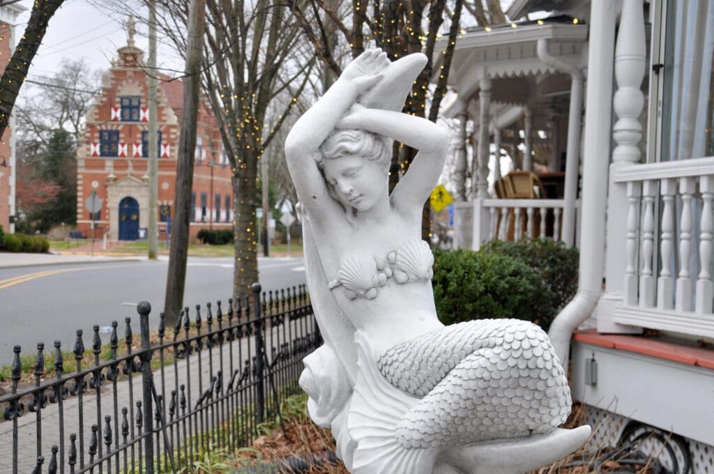 A white mermaid statue sits on a front porch, with her arms raised above her head. Behind her, a wrought iron fence borders a yard and a historic building with a red facade is visible across the street. Trees with string lights line the sidewalk.