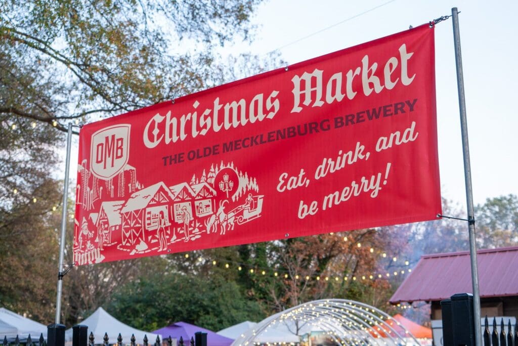 A red banner for Christmas Market at The Olde Mecklenburg Brewery features festive illustrations of a village covered in snow. The text reads Eat, drink, and be merry! with a background of trees and market stalls.