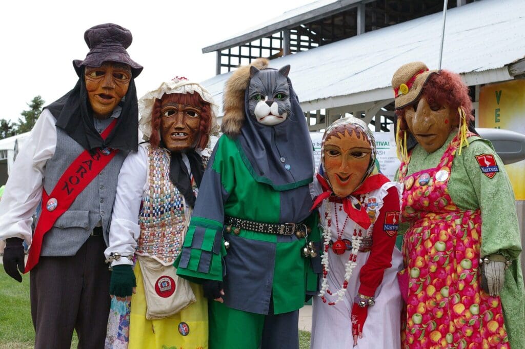 A group of six people dressed in colorful costumes and elaborate masks, including a cat mask, stand together outdoors. They are posing for a photo in front of a building with a sign partly visible in the background.