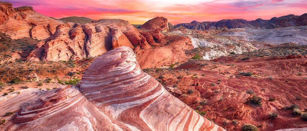 A vibrant landscape of the Valley of Fire State Park at sunset. The scene features red, orange, and white striped rock formations with a colorful sky in the background, blending pink, purple, and yellow hues. Sparse vegetation dots the foreground.