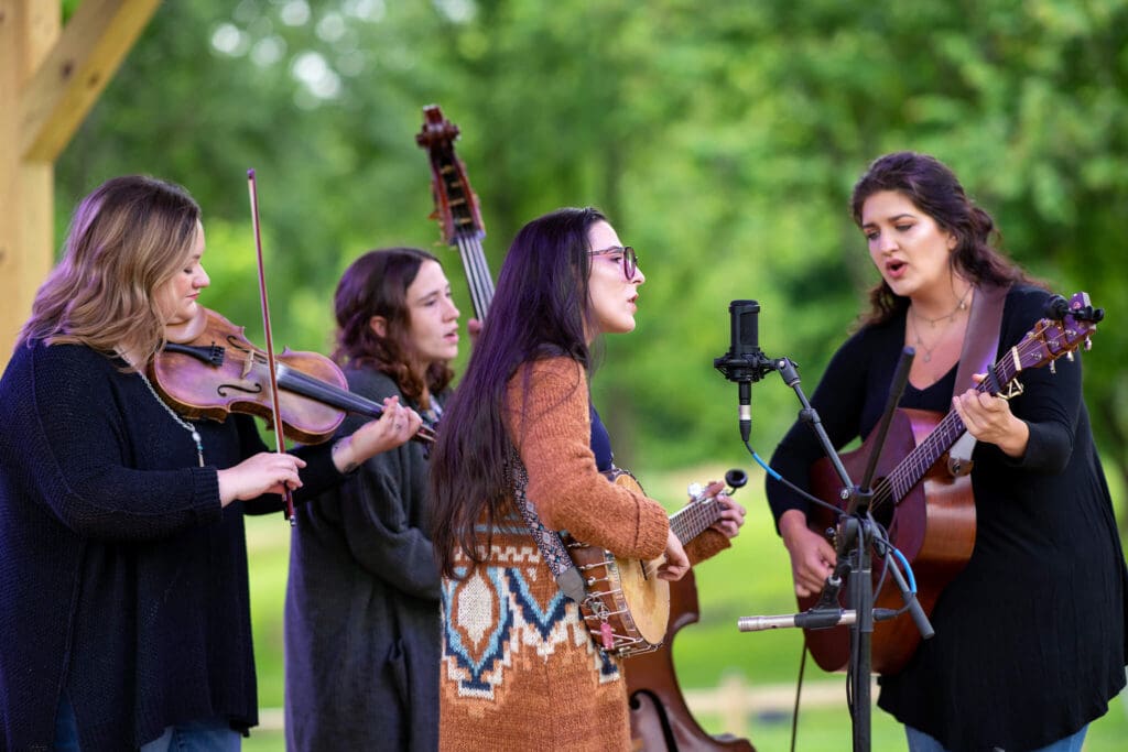 Four women perform music outdoors: one plays a violin, another a cello, the third a banjo, and the fourth a guitar. They sing into a microphone, surrounded by greenery, creating a lively and musical atmosphere.