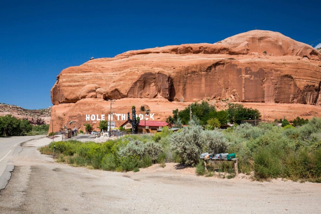 Desert landscape featuring a large red rock formation with Hole N The Rock painted on it. A few small buildings and green shrubs are in the foreground, with a clear blue sky above. A road runs along the left side of the scene.