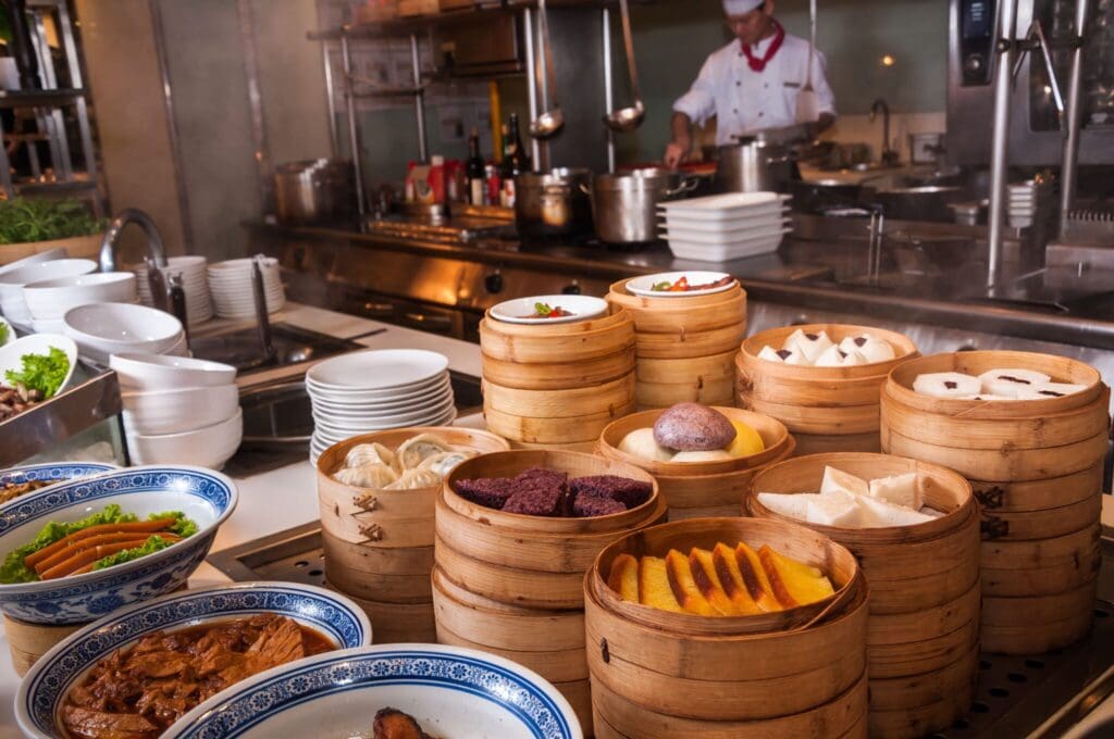 A restaurant kitchen with a chef cooking in the background. In the foreground, there are stacks of bamboo steamers containing various colorful dim sum dishes. Blue and white ceramic plates are also visible, holding more food.
