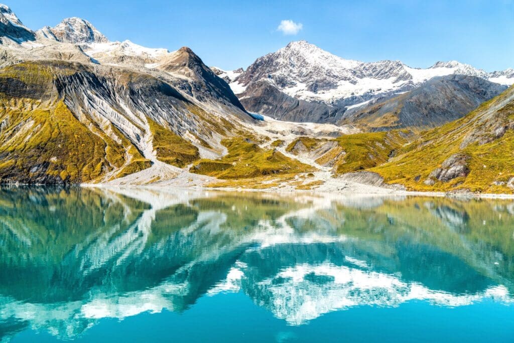 A tranquil lake reflects snow-capped mountains and rugged terrain under a clear blue sky. The landscape features patches of green and yellow vegetation, creating a serene and picturesque natural scene.