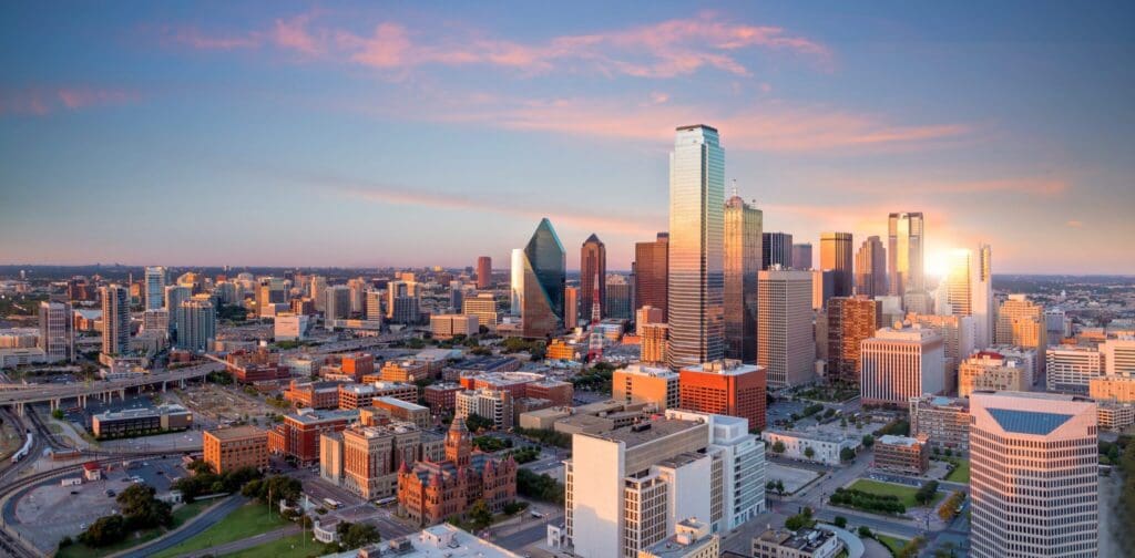 Aerial view of a city skyline at sunset, with a mix of tall modern skyscrapers and smaller buildings. The sky is painted with pink and orange hues. Roads and greenery are visible in the foreground.