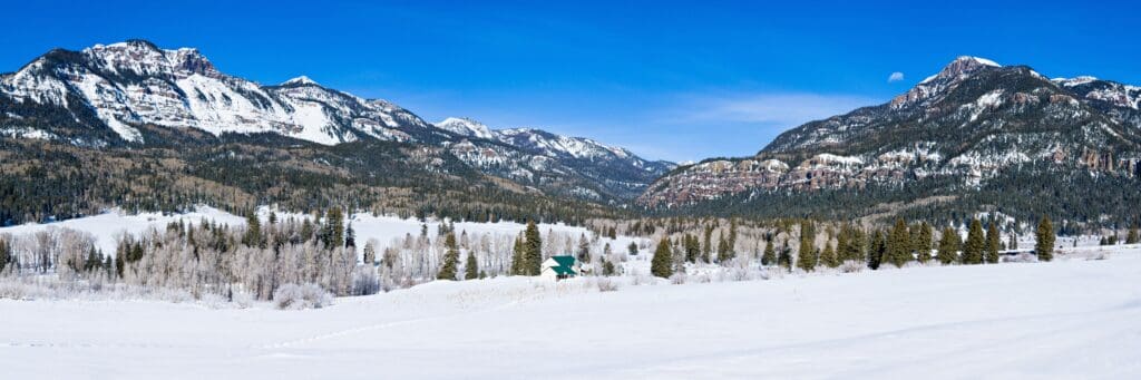 Snow-covered landscape with a cluster of evergreen trees in the foreground and a small cabin. Majestic mountains tower in the background under a clear blue sky. The scene is serene and wintery.