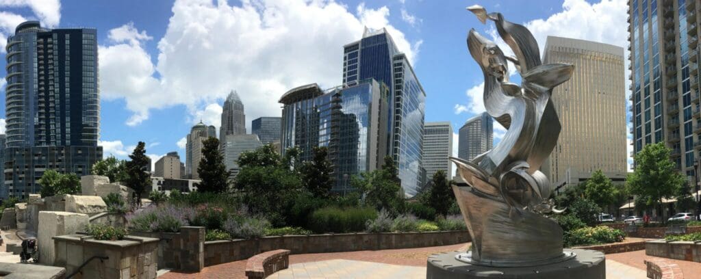 Panoramic view of a modern cityscape with various skyscrapers under a partly cloudy sky. In the foreground, a curving metallic abstract sculpture stands in a landscaped area with flowers and shrubs.