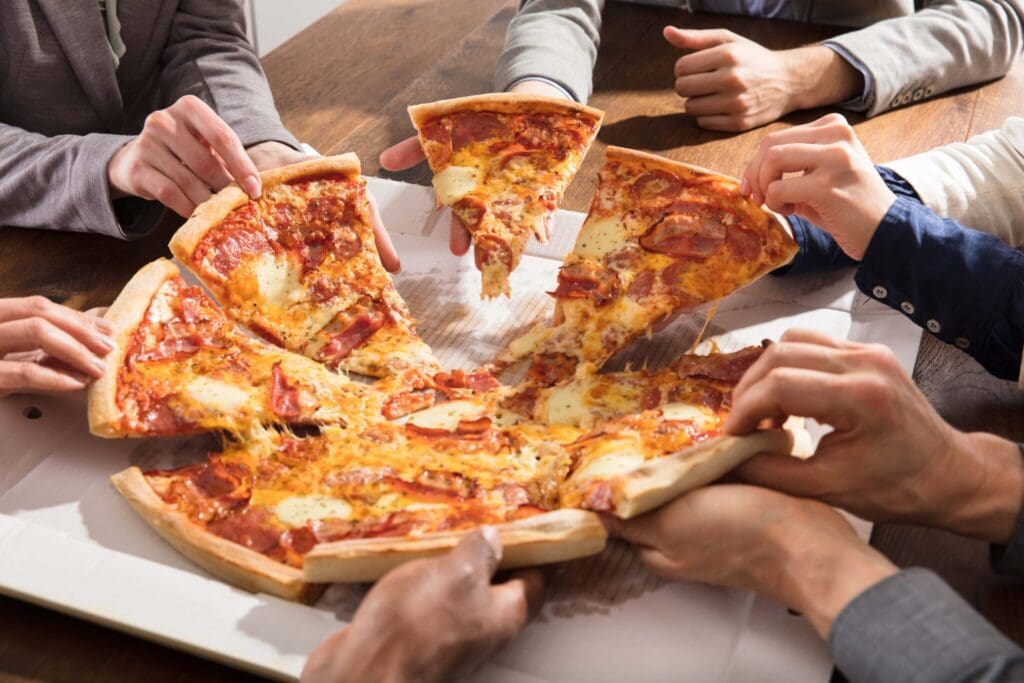 Several hands reaching in to grab slices of a large pepperoni pizza on a wooden table. The pizza is topped with cheese and pepperoni, and is partially cut into slices, with melted cheese stringing between.