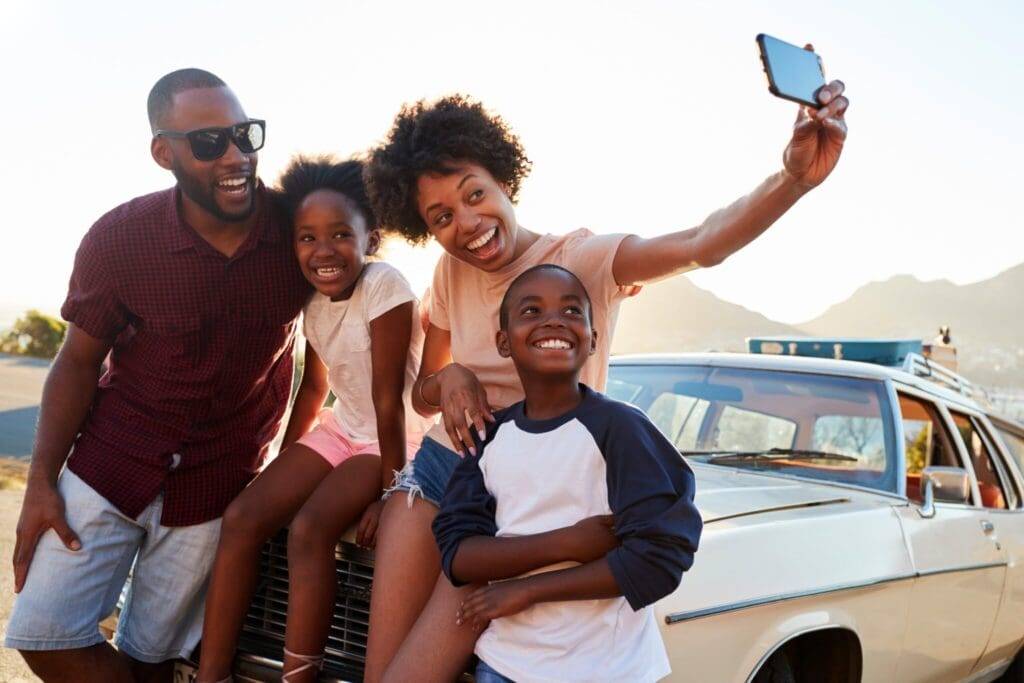 A family of four, including a father, mother, son, and daughter, are joyfully taking a selfie together. They are sitting on the hood of a vintage car with a scenic mountain and sky backdrop during sunset.