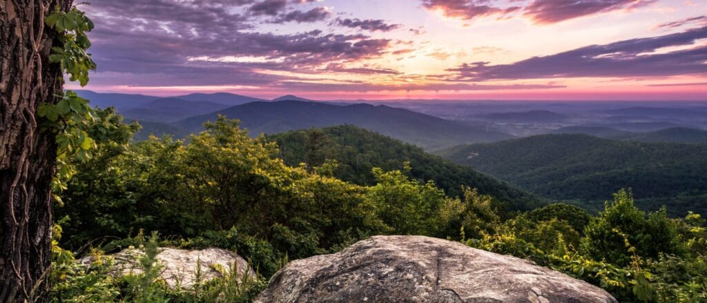 A scenic view of a mountain landscape at sunset. The sky is painted with pink and orange hues, and the sun sets behind distant mountains. Lush green trees and large rocks are in the foreground, creating a serene and picturesque scene.