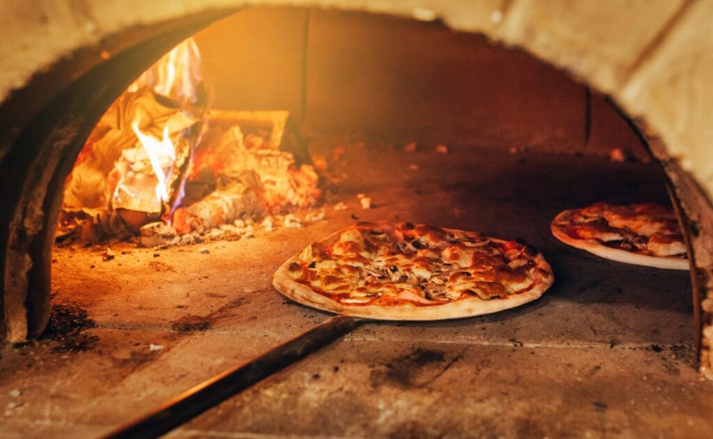 A pizza with a golden crust being baked in a traditional wood-fired oven, with flames visible in the background. Another pizza is partially visible inside the oven.
