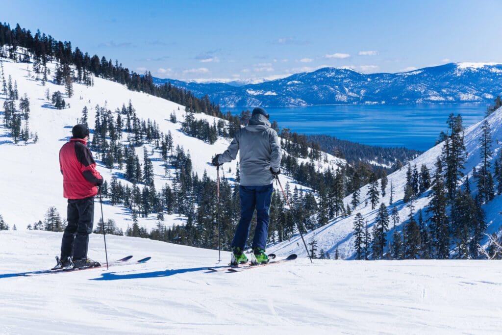 Two skiers stand on a snowy slope, overlooking a scenic view of a lake surrounded by mountains and trees under a clear blue sky. One skier wears a red jacket, the other a gray jacket. Snow-covered peaks are visible in the background.
