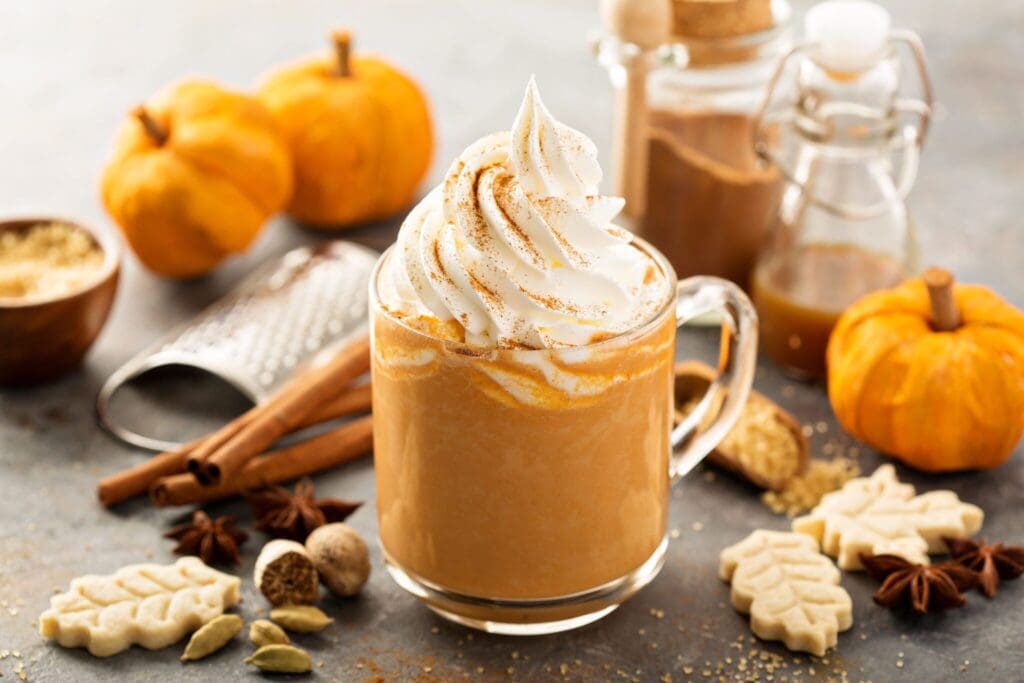 A glass mug filled with whipped cream-topped pumpkin latte, surrounded by small pumpkins, cinnamon sticks, a spice grater, and sugar cookies in leaf shapes, creating an autumn-themed setting.