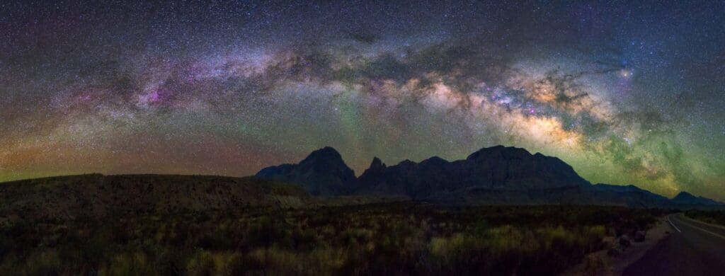 Panoramic view of the Milky Way arching over a dark, mountainous landscape. The sky is filled with vibrant stars and cosmic hues, casting a subtle glow on the rugged terrain below. A quiet road runs along the lower edge of the scene.