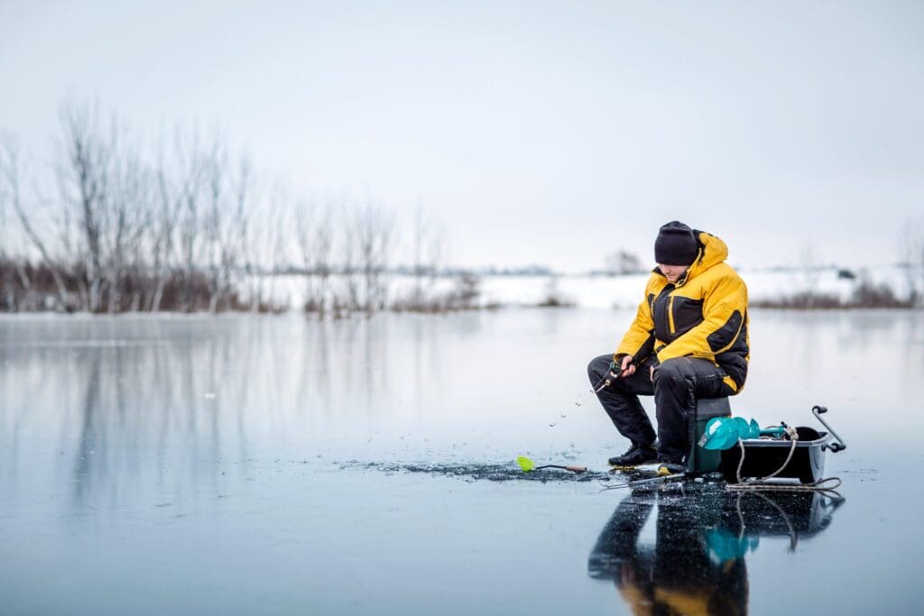 A person sits on a frozen lake ice fishing. They wear a yellow jacket and black pants, holding a fishing rod. A sled with gear is nearby. The background features a serene, snowy landscape with leafless trees under a cloudy sky.