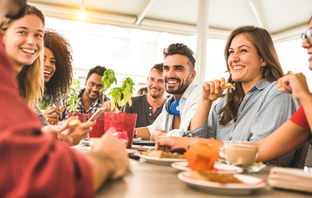 A diverse group of people seated around a table, smiling and laughing. Various drinks and snacks are on the table, creating a warm and friendly atmosphere in a bright, casual setting.