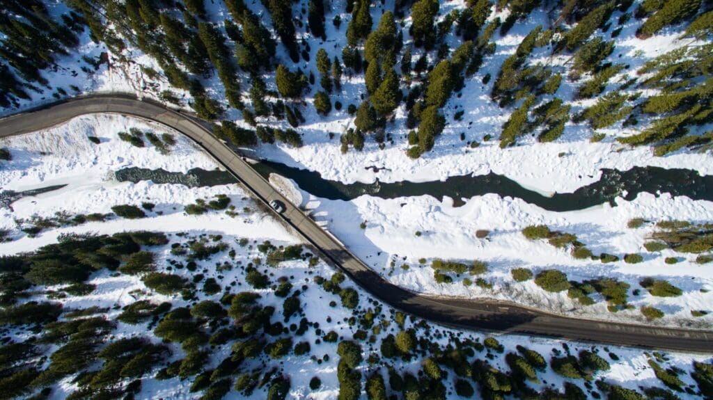 Aerial view of a road winding through a snowy landscape with evergreen trees. A river runs parallel to the road, partially frozen. A single vehicle is traveling along the road, surrounded by the dense forest and snow.
