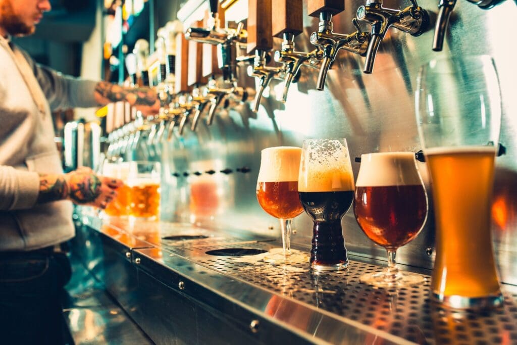 A variety of beers in different glasses sit on a bar counter, partially filled with foam. Behind, a person with tattooed arms operates beer taps, pouring a drink into a mug. The setting appears lively, with a row of taps above the counter.