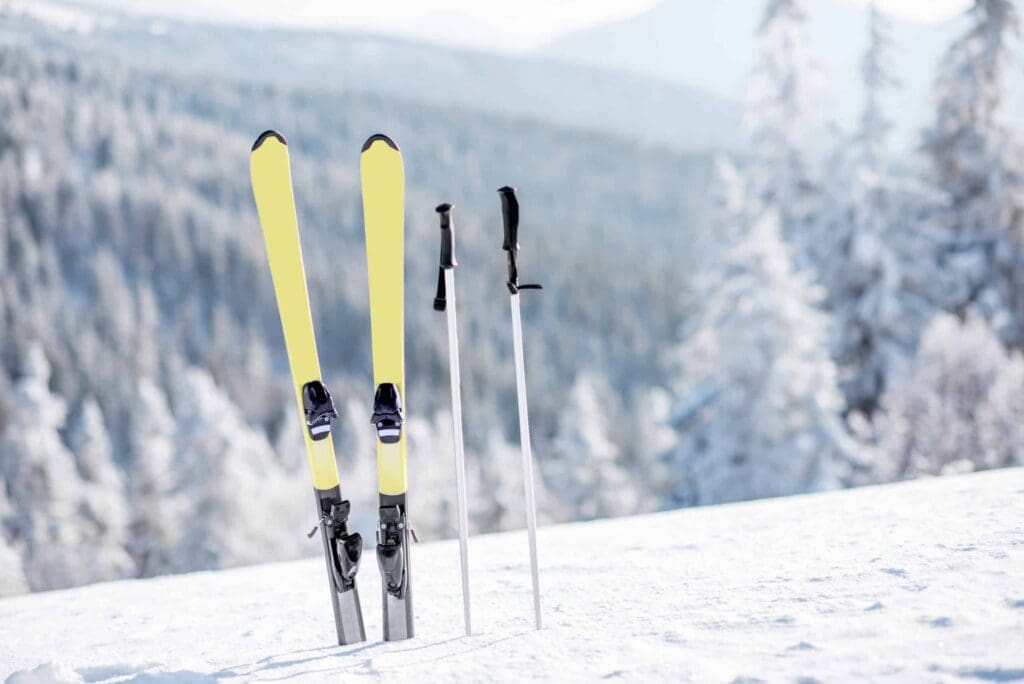 A pair of yellow skis and two ski poles stand upright in the snow against a backdrop of snow-covered trees and a mountainous landscape under a clear sky.