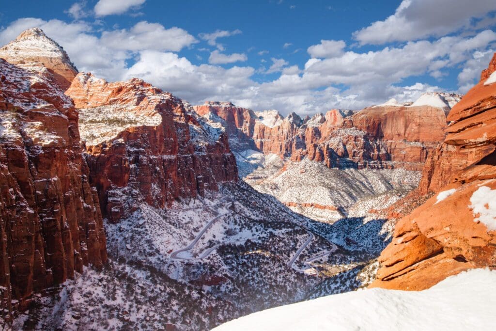 A scenic view of a snow-dusted canyon with towering red rock formations under a partly cloudy blue sky. A winding road is visible at the canyons base, surrounded by snowy patches and vegetation.