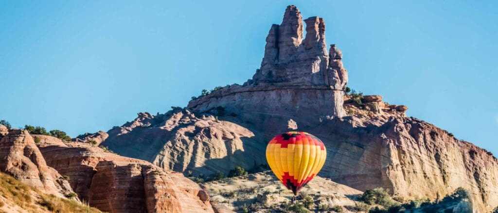 A colorful hot air balloon with a red, yellow, and orange pattern floats in front of towering, rugged rock formations under a clear blue sky. The landscape has a desert-like appearance with sparse vegetation.